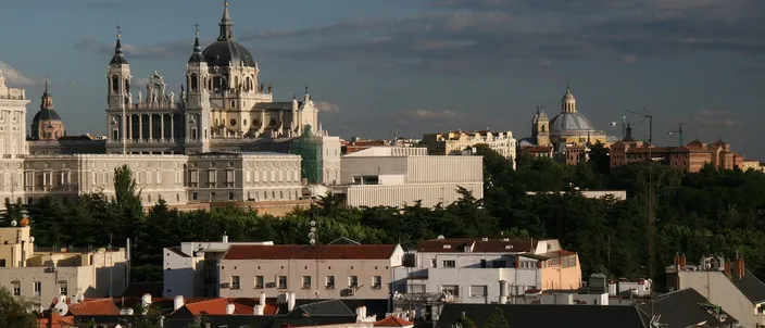 The Almudena Cathedral and Royal Palace rising white above the tiled rooftops of Madrid, seen in warm late-afternoon light from the Templo de Debod hill against a blue sky with scattered cloud.