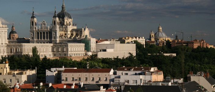 The Almudena Cathedral and Royal Palace rising white above the tiled rooftops of Madrid, seen in warm late-afternoon light from the Templo de Debod hill against a blue sky with scattered cloud.