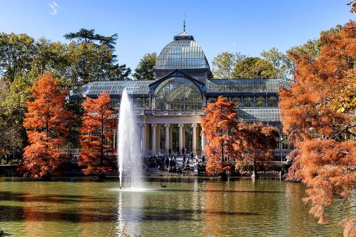 A glass and iron pavilion with an arched dome on a brick base, standing behind a pond with a tall jet of water in front of it, rust-orange cypresses lining both banks under a clear blue sky.