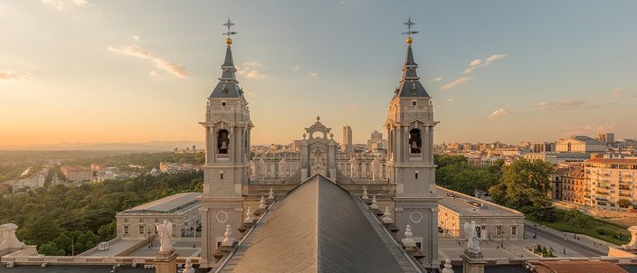 Two stone bell towers with clock faces seen from above and behind, a tiled roof ridge running between them, the low rooftops of a city and a wooded hillside stretching to the horizon under a warm orange sky.