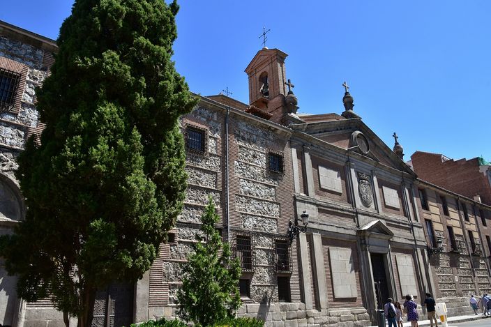 The austere stone and brick side of the Descalzas Reales convent church rising straight off a narrow Madrid street, a small bell gable and cross above the roofline, a dark cypress in front of it and a few people at the doorway in strong sun.