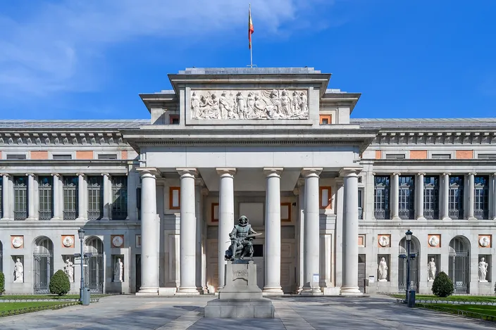 The Velázquez front of the Prado Museum in full sun, a colonnaded portico under a carved relief pediment, a seated bronze statue of the painter on a plinth at the foot of the steps, blue sky behind.