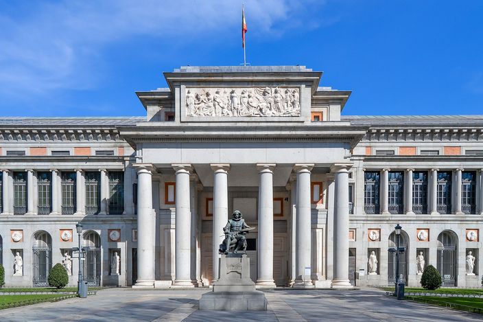 The Velázquez front of the Prado Museum in full sun, a colonnaded portico under a carved relief pediment, a seated bronze statue of the painter on a plinth at the foot of the steps, blue sky behind.