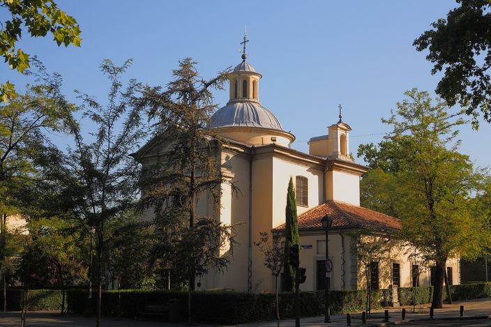 A small domed neoclassical chapel with pale rendered walls and a slate dome, a lantern and cross above it, standing among trees behind a low railing in warm late-afternoon light.