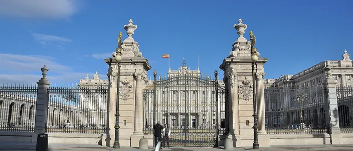 The Royal Palace of Madrid seen head-on through the open ironwork gates of a wide paved forecourt, two tall stone piers topped with carved urns framing the long white facade under a deep blue sky.