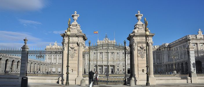 The Royal Palace of Madrid seen head-on through the open ironwork gates of a wide paved forecourt, two tall stone piers topped with carved urns framing the long white facade under a deep blue sky.