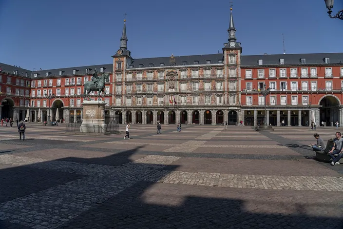 Plaza Mayor's arcaded red-brick facade with twin spires and the bronze equestrian statue of Philip III on the wide paved square under a clear blue sky