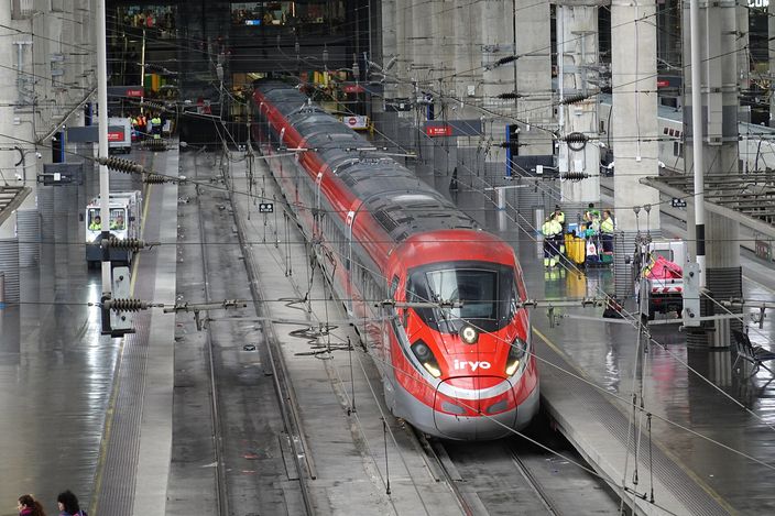 A red and white Iryo high-speed train stands at a platform inside Madrid Puerta de Atocha station, under the vast steel-and-glass trainshed with overhead catenary wires and reflective wet tracks.