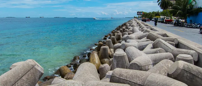 Malé's western seafront: gray concrete tetrapods of the breakwater piled along the shore, clear turquoise water stretching to the horizon on the left, and a seafront road with parked scooters, palms and apartment blocks on the right.