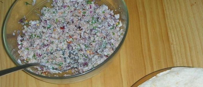 A glass bowl of mas huni — shredded tuna worked with grated coconut, red onion and chilli — with a stack of thin roshi flatbread beside it on a wooden table.