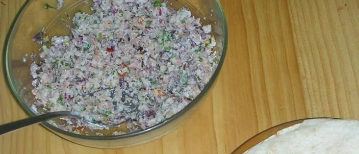 A glass bowl of mas huni — shredded tuna worked with grated coconut, red onion and chili — with a stack of thin roshi flatbread beside it on a wooden table.