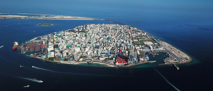 Aerial view of Malé, a densely packed island city crammed with high-rise apartment blocks and government buildings edge to edge, ringed by a harbour and turquoise sea, with a cargo ship and small boats around its shores.