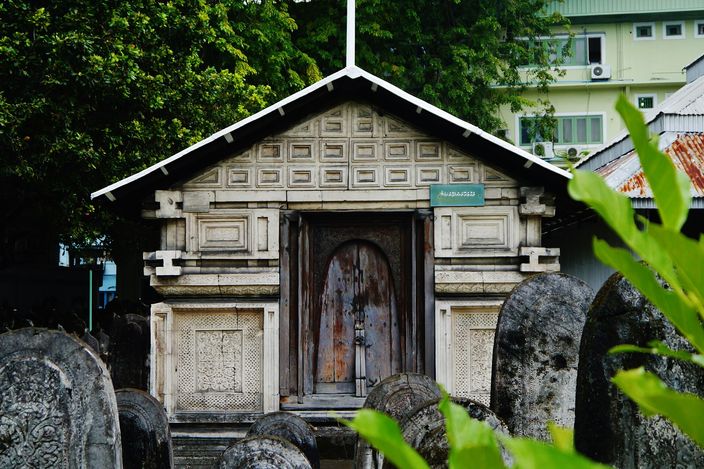 Weathered coral-stone gravestones carved in fine relief in the old graveyard beside Hukuru Miskiy, with the decorated stone facade of a shrine behind them.