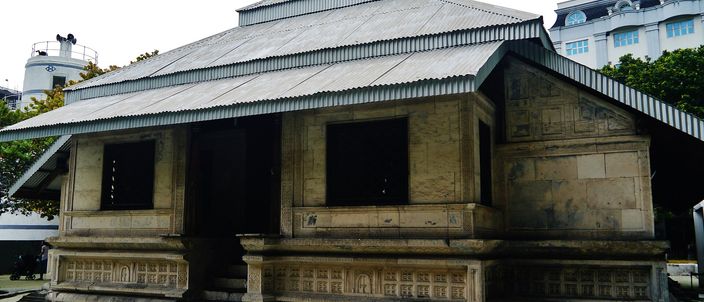 Hukuru Miskiy, Malé's old Friday mosque: a low coral-stone building with finely carved walls under a pitched corrugated roof, modern city buildings rising behind it.
