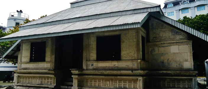 Hukuru Miskiy, Malé's old Friday mosque: a low coral-stone building with finely carved walls under a pitched corrugated roof, modern city buildings rising behind it.