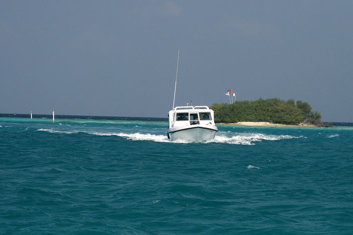 A small white motorboat crossing turquoise lagoon water at speed, throwing up a wake, with a low palm-fringed island in the background.
