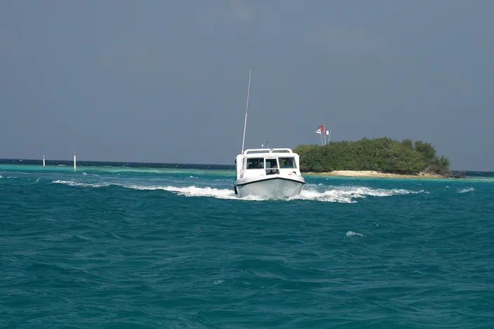A small white motorboat crossing turquoise lagoon water at speed, throwing up a wake, with a low palm-fringed island in the background.