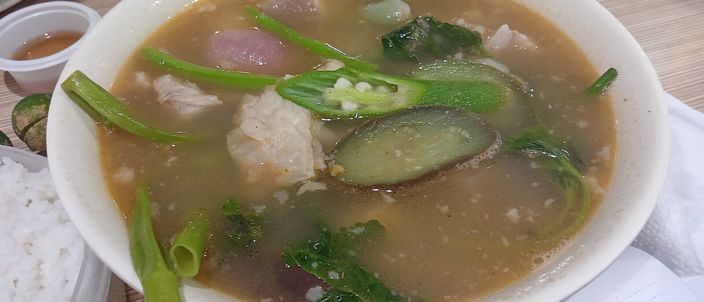 A bowl of sinigang na baboy — pork in a cloudy, sour tamarind broth crowded with taro, aubergine, long beans and greens, served with rice alongside.