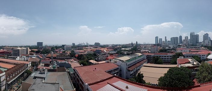 Panoramic rooftop view over the red-tiled roofs of Intramuros, the walled Spanish old town, with the modern Manila skyline rising in the distance under a bright cloudy sky