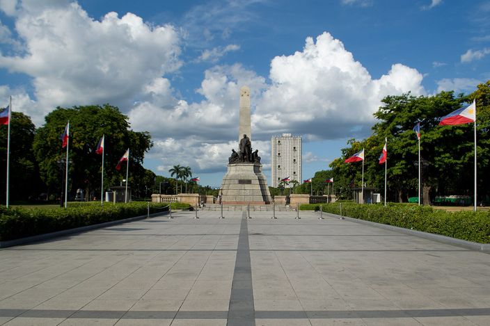 A tall stone monument with an obelisk and a group of bronze figures at its base, standing at the end of a wide empty paved plaza lined with national flags on poles, trees and a mid-rise building behind under a blue sky with white cumulus.