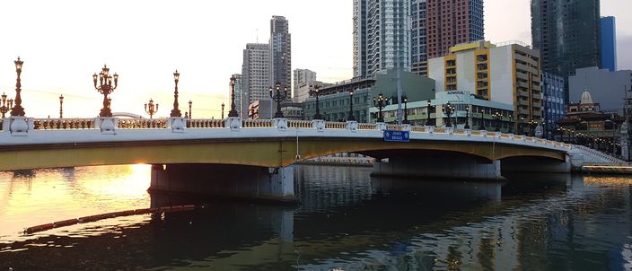 A low road bridge with ornate gilded lamp posts and white balustrades spanning a wide river at sunset, high-rise towers and older commercial buildings along the far bank, the orange sky reflected in the water.