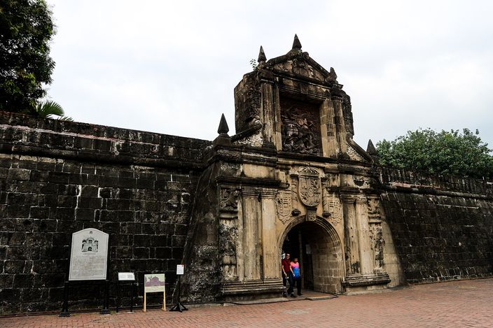The carved stone gateway of Fort Santiago set into the dark rampart wall of Intramuros, its pediment topped with obelisk finials above a relief and coat of arms, two visitors standing in the arched opening, a cast historical marker on the wall to the left