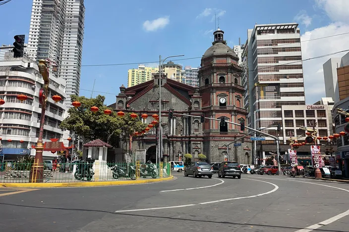 Binondo Church seen from a street junction in Chinatown, its octagonal tiered bell tower and stone dome rising above the facade under a blue sky, rows of red paper lanterns strung across the road in front of it