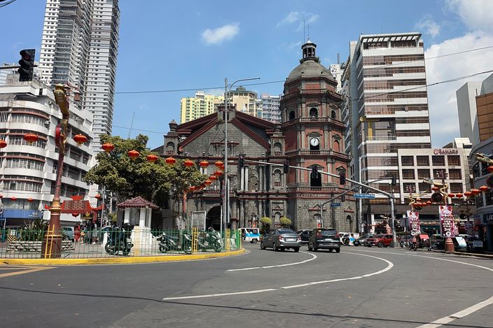 Binondo Church seen from a street junction in Chinatown, its octagonal tiered bell tower and stone dome rising above the facade under a blue sky, rows of red paper lanterns strung across the road in front of it