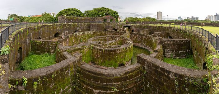 Wide view down into the moss-covered stone bastion of Baluarte de San Diego in Intramuros, its circular keep and radiating ramparts open to the sky with grass growing between the walls, visitors on the outer rampart and modern towers on the skyline behind