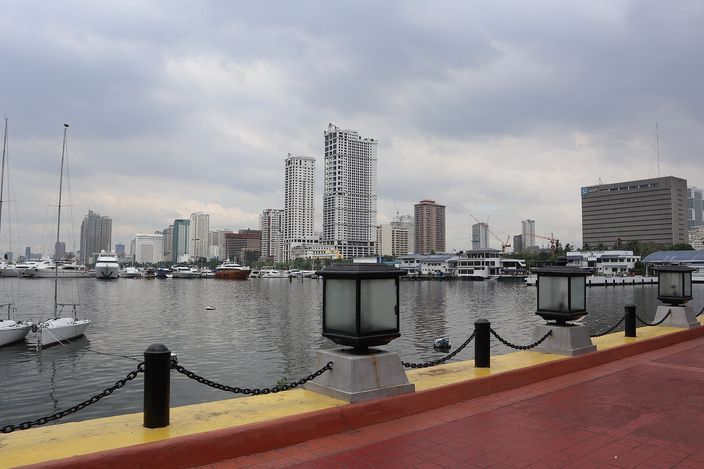 View across the Manila Bay marina from Harbour Square promenade, yachts moored at left, a curving line of lamp posts and chain railing along a red-and-yellow quay, high-rise towers on the far shore under a grey overcast sky