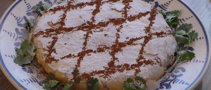 A round Moroccan chicken pastilla dusted with powdered sugar and marked with a lattice of cinnamon, garnished with fresh cilantro on a blue-patterned plate