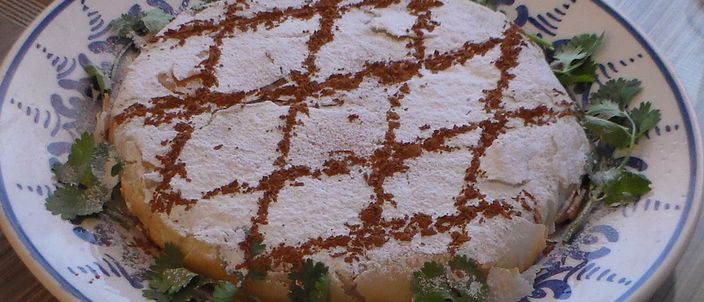 A round Moroccan chicken pastilla dusted with icing sugar and marked with a lattice of cinnamon, garnished with fresh coriander on a blue-patterned plate