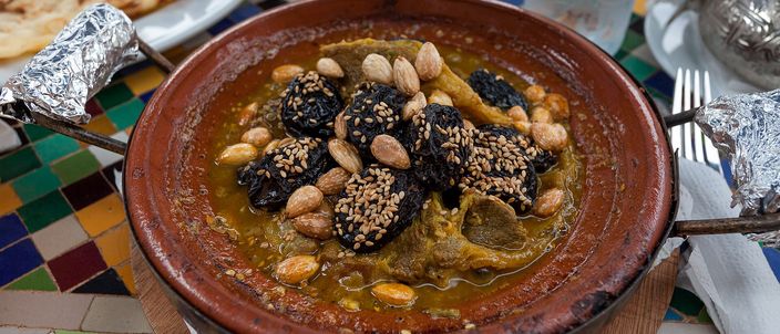 A Moroccan lamb tagine with prunes, whole almonds and sesame seeds in a plain unglazed terracotta cooking pot, set on a colourful zellige-tiled table with bread alongside