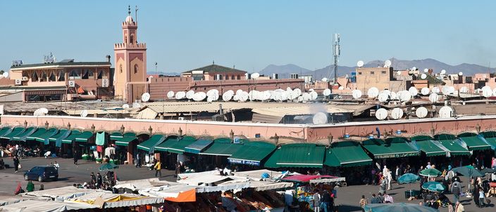 The open square of Jemaa el-Fna at midday, rows of stalls under green and white awnings, low ochre buildings and a minaret behind them, and the High Atlas on the horizon under a clear sky