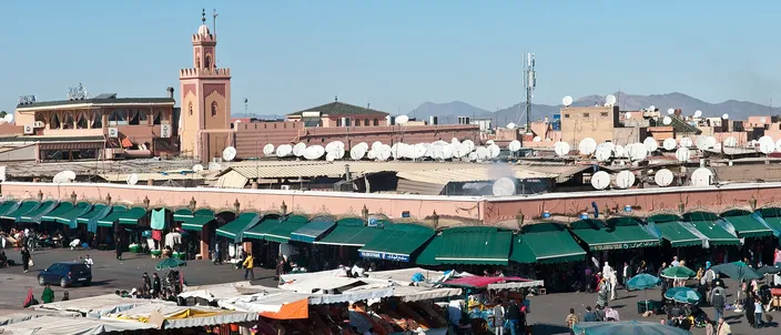 The open square of Jemaa el-Fna at midday, rows of stalls under green and white awnings, low ochre buildings and a minaret behind them, and the High Atlas on the horizon under a clear sky