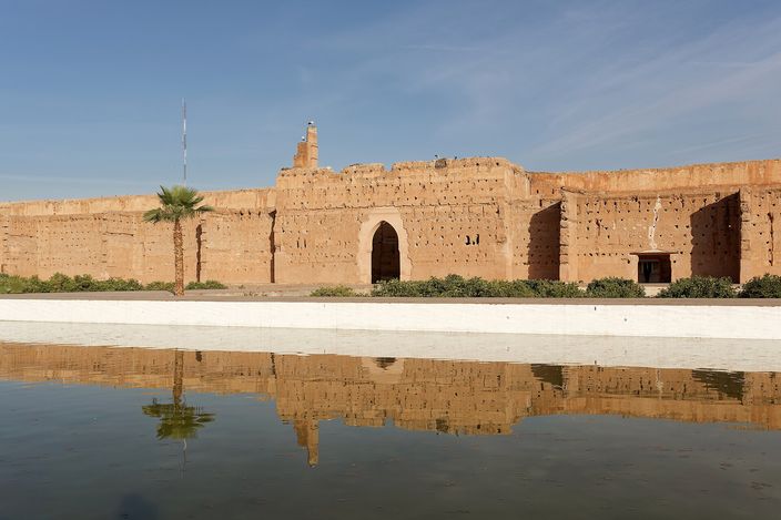 The long earth-red outer wall of El Badi Palace with its tall arched gateway and a single palm, mirrored in the still rectangular pool of the sunken courtyard under a clear blue sky