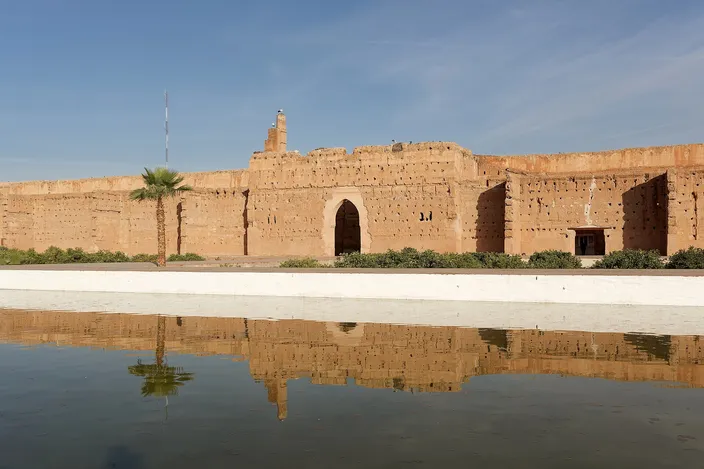 The long earth-red outer wall of El Badi Palace with its tall arched gateway and a single palm, mirrored in the still rectangular pool of the sunken courtyard under a clear blue sky