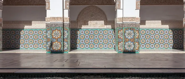 Three tall arched bays of the main courtyard of the Ben Youssef Madrasa in Marrakesh, their piers and dados faced in blue, green and ochre zellij mosaic beneath panels of finely carved white stucco and a dark carved cedar cornice, standing above a polished marble floor with the edge of the courtyard's water along the bottom of the frame