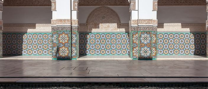Three tall arched bays of the main courtyard of the Ben Youssef Madrasa in Marrakesh, their piers and dados faced in blue, green and ochre zellij mosaic beneath panels of finely carved white stucco and a dark carved cedar cornice, standing above a polished marble floor with the edge of the courtyard's water along the bottom of the frame