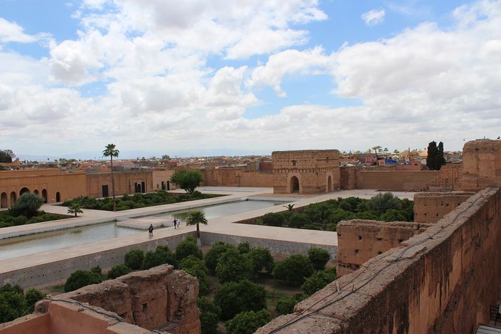 Looking down from the ramparts of El Badi Palace over its sunken courtyard: two long rectangular pools and sunken groves of orange trees inside high bare earth walls, with two people walking on the path between them and the low roofs and palms of Marrakesh beyond, under broken cloud.
