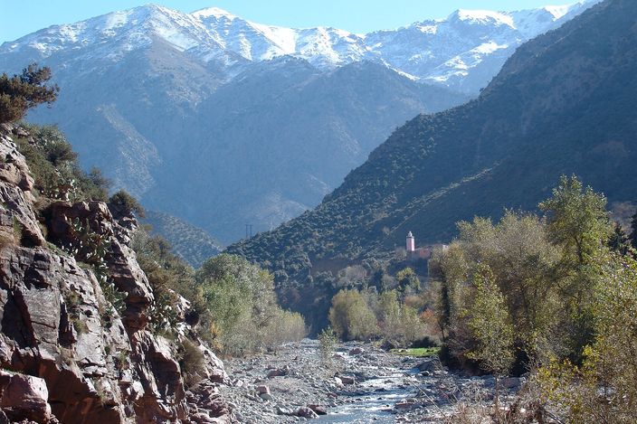 The Ourika valley: a wide stony river bed running between red rock and stands of green trees, a village with a minaret on the far bank, and the snow-covered ridge of the High Atlas closing the head of the valley under a clear sky.