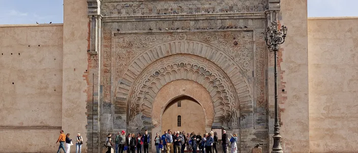 The Almohad gateway of Bab Agnaou seen head on: a tall pointed horseshoe arch set inside a rectangular stone frame of carved concentric bands and floral corner medallions, cut into a long pink-ochre rampart wall, with a small group of people standing at its foot beside a zebra crossing.