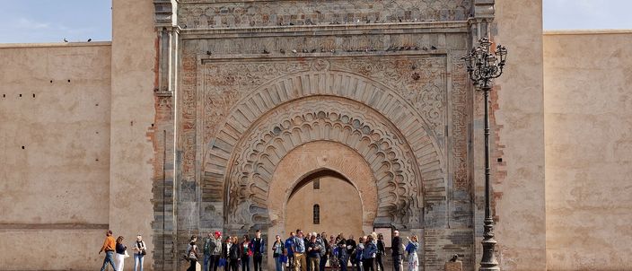 The Almohad gateway of Bab Agnaou seen head on: a tall pointed horseshoe arch set inside a rectangular stone frame of carved concentric bands and floral corner medallions, cut into a long pink-ochre rampart wall, with a small group of people standing at its foot beside a zebra crossing.