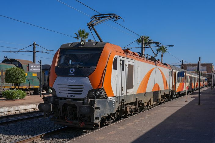 An orange and white ONCF electric locomotive standing at a platform of Marrakesh railway station in bright sun, palms behind it and overhead catenary above the tracks