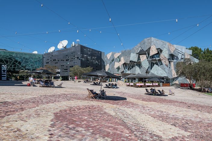 Federation Square in Melbourne on a clear day: angled zinc, sandstone and glass facades across the far side, a sloping cobbled forecourt in the foreground, and scattered deckchairs and umbrellas with a few people sitting out.