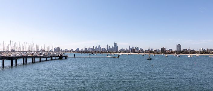 A wide view out over Port Phillip Bay from St Kilda, with a long low jetty and a marina full of yacht masts at the left, small boats at anchor on flat blue water, and the low Melbourne CBD towers on the far shore under a clear sky.