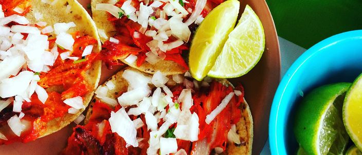 Three tacos al pastor on small corn tortillas, the adobo-red shaved pork heaped with chopped white onion, with lime wedges and a bowl of cut limes alongside.