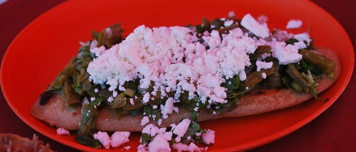 A single oval tlacoyo on a red plate, split along the top and dressed with griddled nopales, shredded meat and crumbled white cheese.