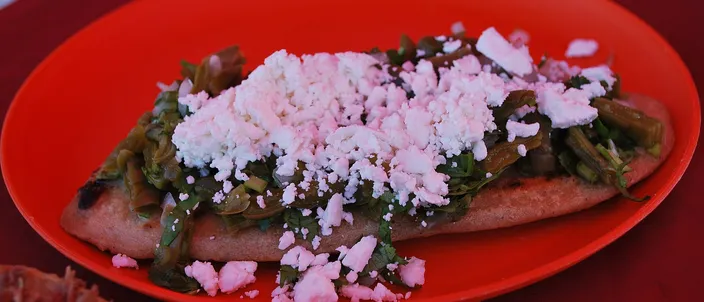 A single oval tlacoyo on a red plate, split along the top and dressed with griddled nopales, shredded meat and crumbled white cheese.