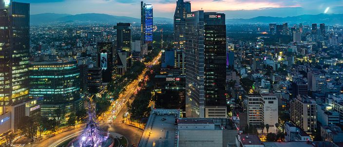 Dusk aerial along Paseo de la Reforma, the Angel of Independence roundabout glowing at lower left, glass towers lining the avenue and mountains on the horizon under a blue-gold sky